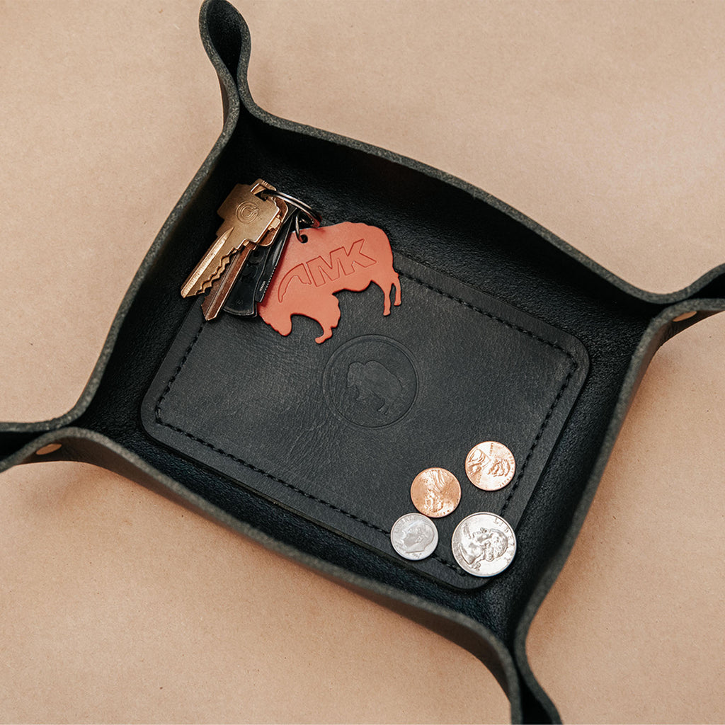 An overhead view of the black leather valet tray handmade by local leatherworking company ColsenKeane. These trays are designed to keep small items organized and are stamped in the center with the Mountain Khakis signature bison logo.